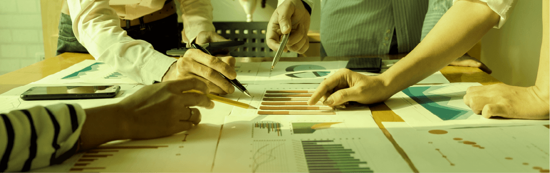 The hands of sales professionals all learn toward graphs and charts in the center of a table. The sepia-like tones indicate that this is an old way of doing things.