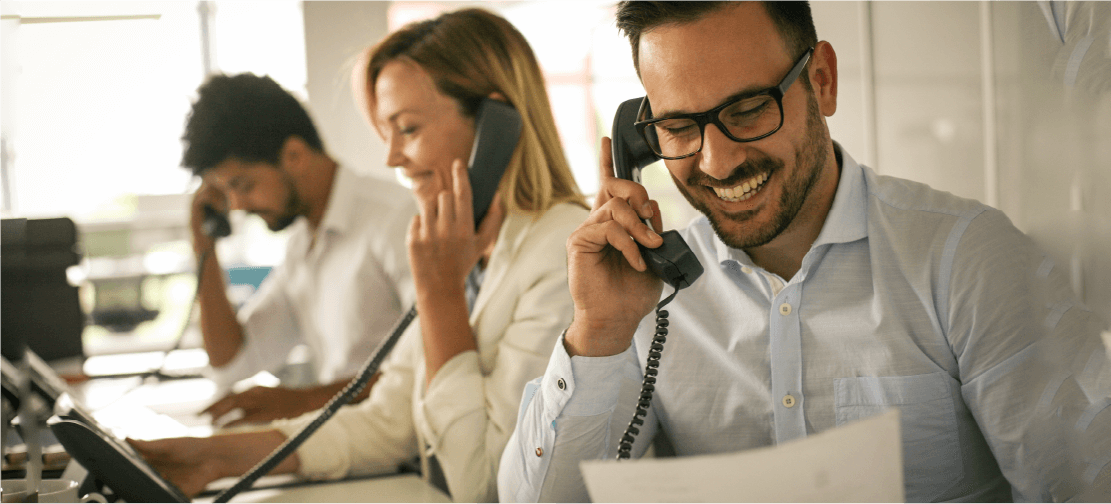 Young, happy sales professionals sit lined up at a busy table, all answering phone calls with their customers.